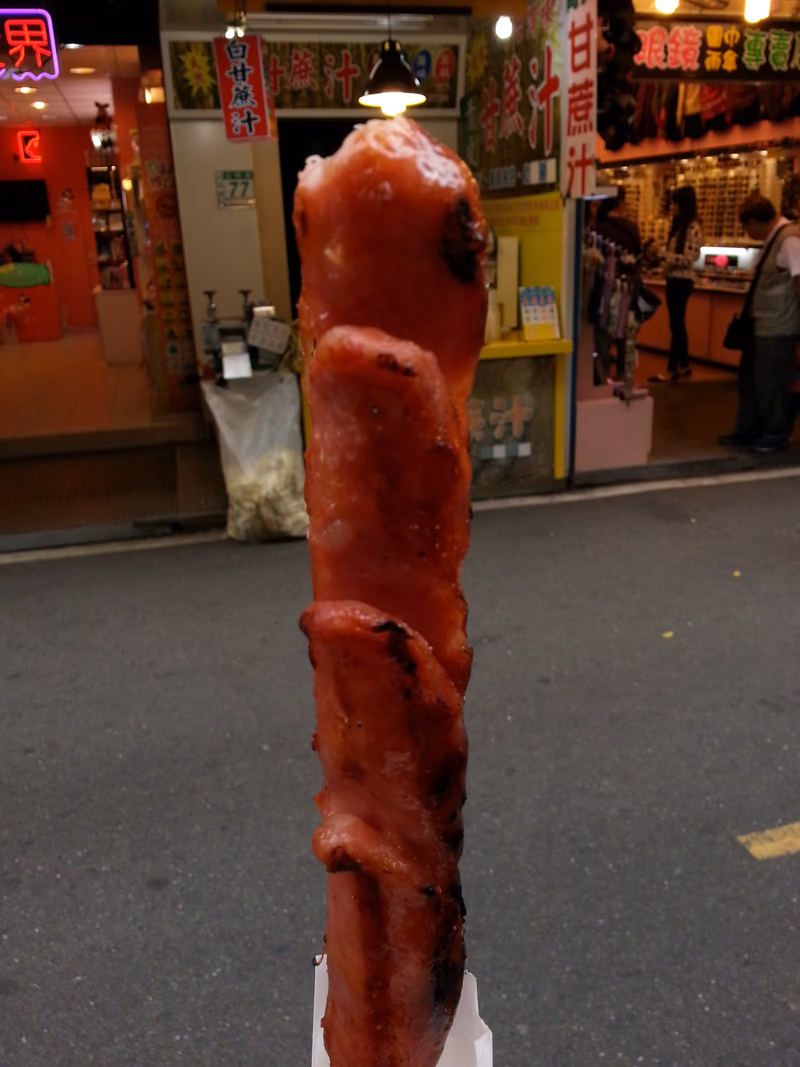 A close-up of a food stand with a large, red, grilled meat-like food item in the foreground.